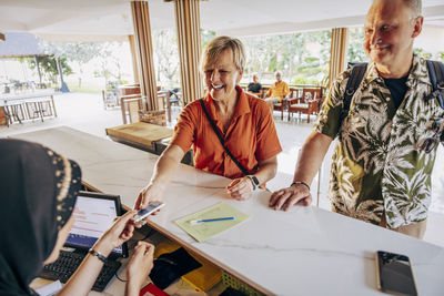 Happy senior woman taking room key from female receptionist at desk in hotel