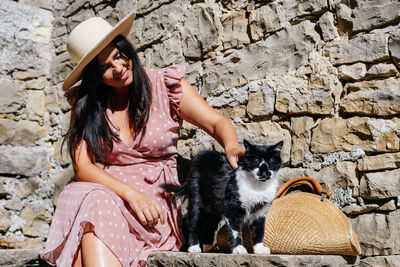 Young woman in pink dress sitting on stone bench in old town, petting adorable cat.