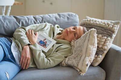 Sad mature woman holding family photo while lying with closed eyes on sofa at home