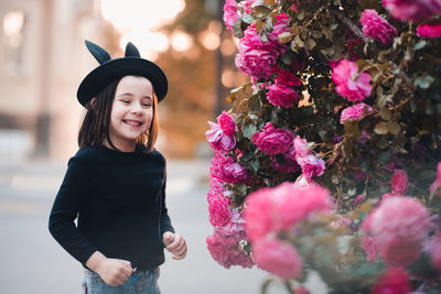 Cute stylish child girl 5-6 year old wear hat and black top posing over rose flower bushes outdoors.