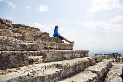 Man in a blue linen shirt and shorts sits on an old stone staircase  colosseum in pamukkale turkey