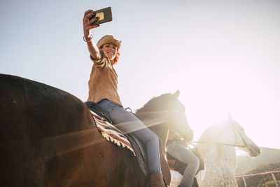 Low angle view of young woman taking selfie while riding horse against clear sky