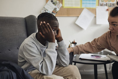 Non-binary mental health professional consoling sad male student sitting on chair in office