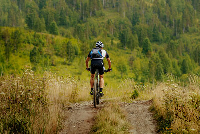 Back male cyclist with backpack on mountain bike riding trail in grass field