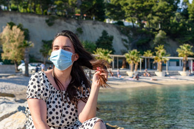 Young woman wearing surgical mask, sitting and relaxing on beach in summer.