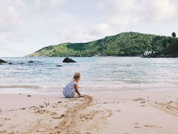 Rear view of girl sitting on beach against mountain