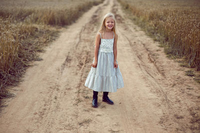 Blonde girl stands on an earthen path in a long dress among a wheat field in the summer