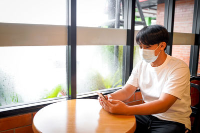 A handsome young teenage boy wear mask playing on smart mobile phone waiting in cafeteria. 