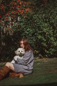 Portrait of young woman holding dog while sitting on grassy field against trees