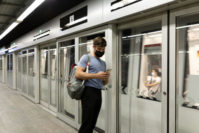 Young male commuter using smart phone while standing by moving train at subway station during covid-19