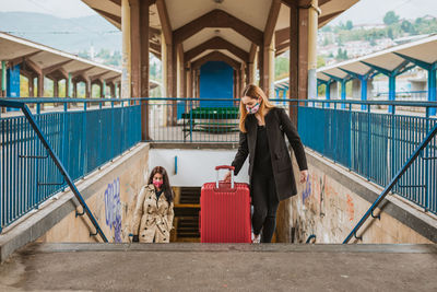 Young women wearing mask holding suitcase standing on staircase at railroad platform