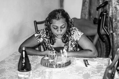 Young woman blowing candles on birthday cake at table in home