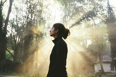 Side view of young woman standing against trees during sunrise
