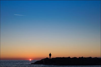 Silhouette man standing on rocks by sea against clear sky during sunset