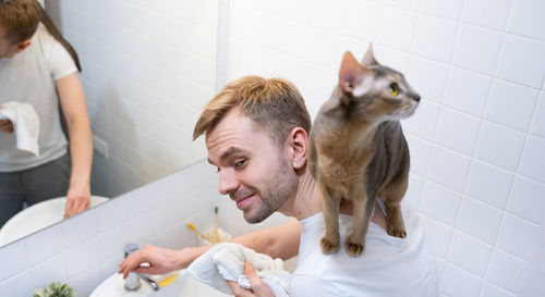 A young smiling blond man is washing his face with a abyssinian cat on his shoulders.domestic life