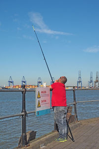 Rear view of man fishing by sea against sky