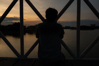 Rear view of silhouette man standing by railing against sky during sunset