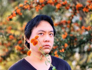 Close-up of young asian man holding orange mums flowers against orange rowan berry trees.