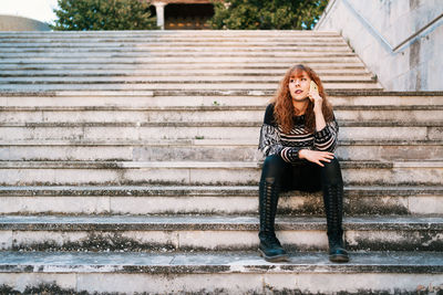 Young red-haired caucasian woman talking on the phone sitting on the stairs outside