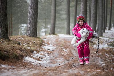 Girl in warm clothing carrying toy tiger while walking in forest during winter