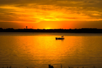 Silhouette people on boat in lake against orange sky