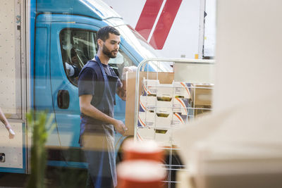 Young male owner standing by rack against food truck seen through restaurant window glass