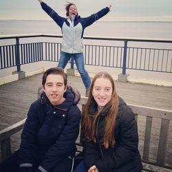 Portrait of siblings sitting on bench with cheerful mother in background at pier