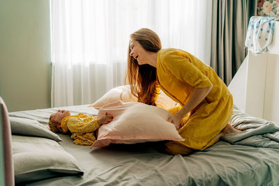 Laughing mother fights pillows with her little daughter on the bed.