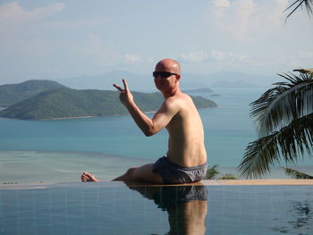 Portrait of smiling mature man showing peace sign at infinity pool against sea