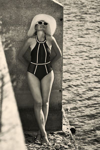 Portrait of a woman in a swimsuit, hat and sunglasses in summer on the riverbank by a concrete wall