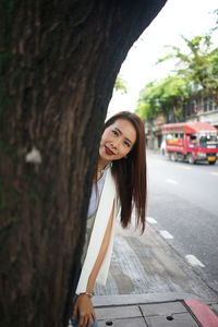 Portrait of smiling woman standing behind tree on sidewalk