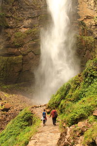 Rear view full length of man and woman holding hands against waterfall