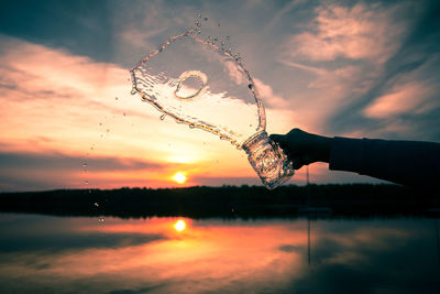 Cropped hand pouring water from jar against sky during sunset