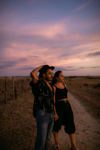 Rear view of couple standing on field against sky during sunset