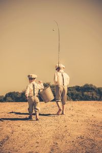 Rear view of siblings with bucket and fishing rod walking on field against sky