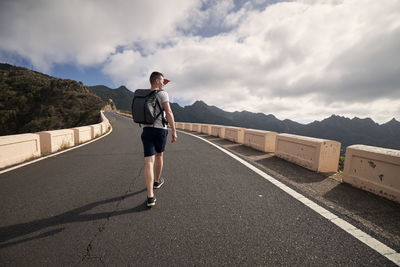 Rear view of man with backpack on mountain road. young tourist enjoys trip on sunny day. 