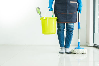 Low section of person holding mop and bucket against wall on floor at home