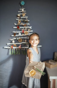 Portrait of smiling girl holding gift against christmas tree hanging on wall