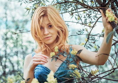 Young blonde woman in a denim top stands among flowering branches in nature and looks at the camera.