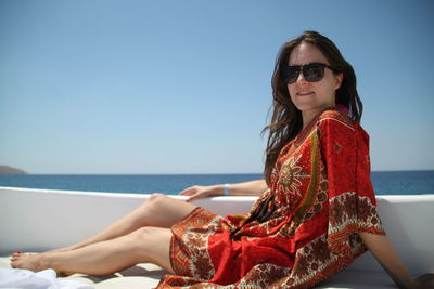 Young woman wearing sunglasses sitting on boat deck in sea against clear sky