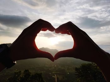 Cropped image of hand holding heart shape against sky during sunset