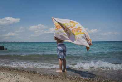 Low section of boy holding towel while standing on shore at beach against sky