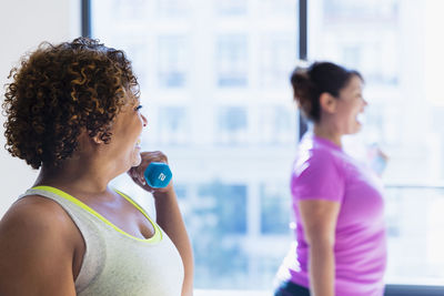 Happy friends lifting dumbbells while exercising against window in yoga studio