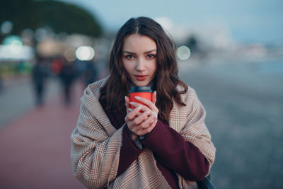 Portrait of beautiful young woman holding smart phone while standing outdoors