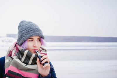 Close-up of young woman smoking while standing on snow covered field against sky