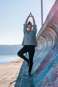 Full length of young woman doing yoga by wall against clear sky