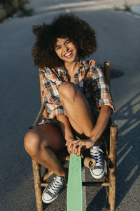 Happy black female with in trendy summer outfit and with afro hairstyle sitting on chair on asphalt road with green penny board and looking at camera