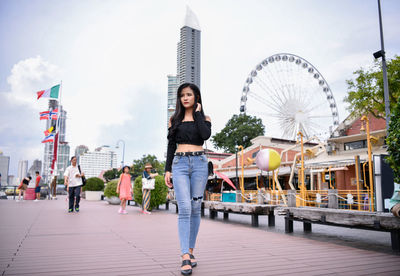 Young woman walking on footpath against ferris wheel and sky in city