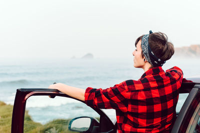 Back view of tranquil female hipster with piercing in casual shirt and bandana standing near car and looking away at empty seaside