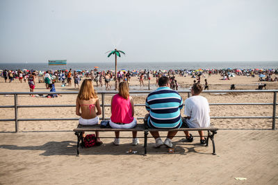 Rear view of people sitting on bench at beach against sky